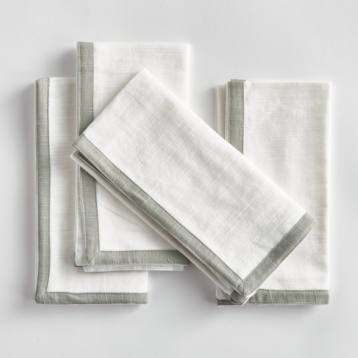 White and sage dinner napkins, folded in a layered arrangement, showing the border and square cotton fabric. Transitional