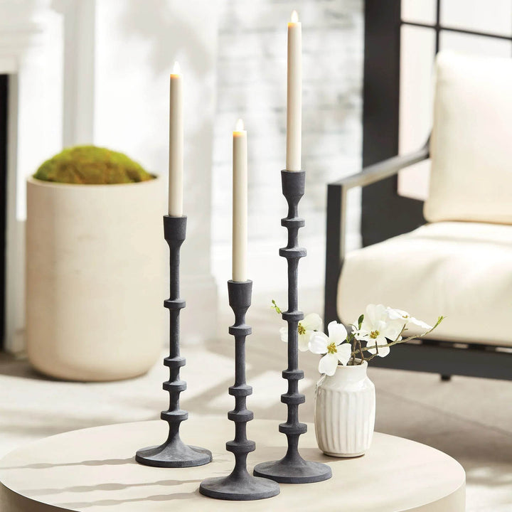 Trio of industrial black taper holders styled on round table with flowers and seating in background – Transitional