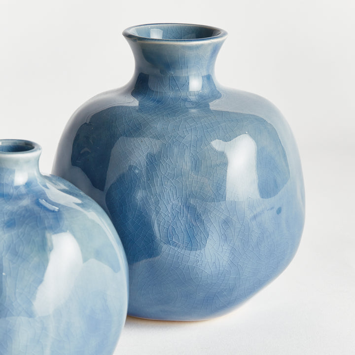 Close view of a blue ceramic bud vase with mottled glaze and an uneven organic surface, beside its small match. Transitional