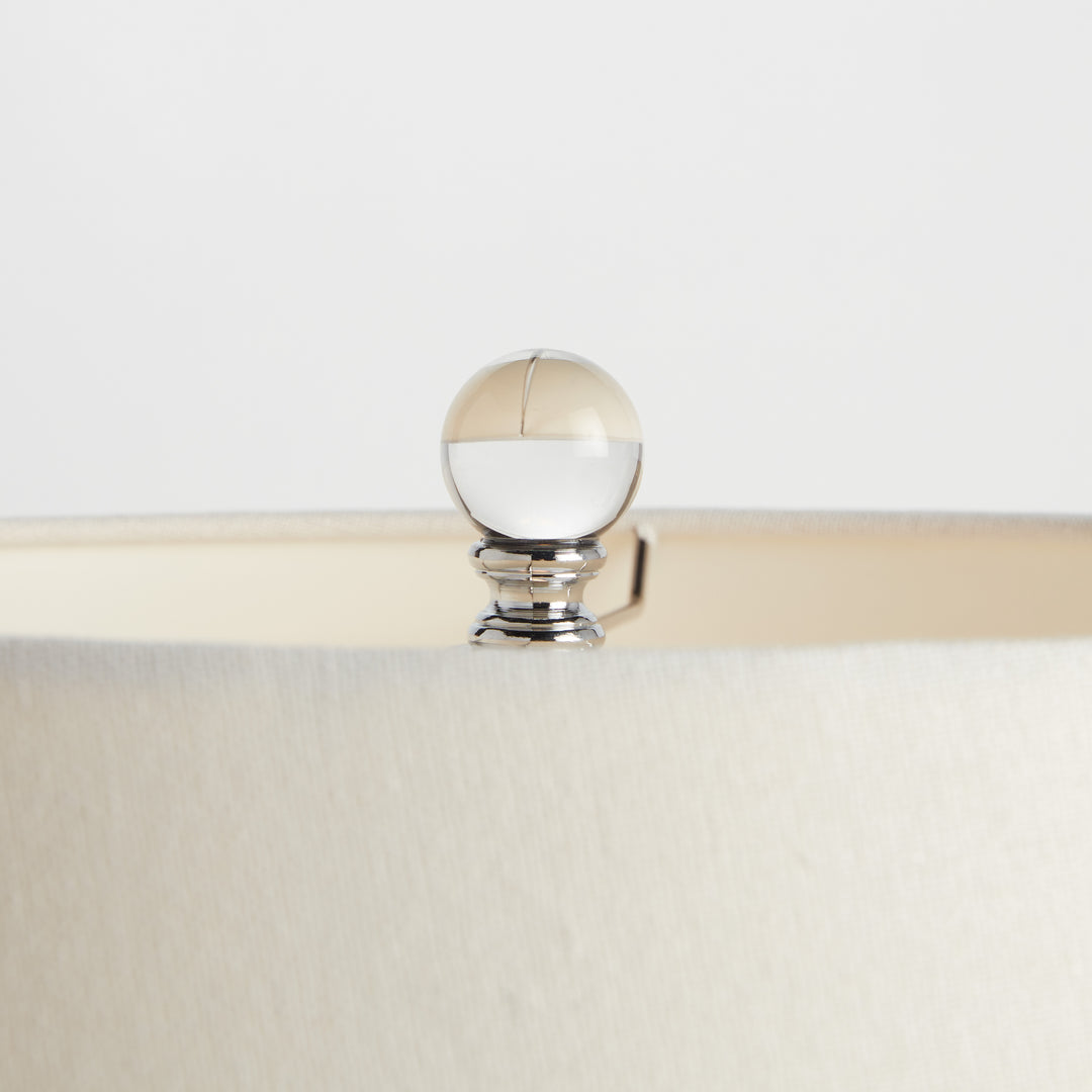 Faceted crystal finial on top of white linen lamp shade with polished silver-tone fitting detail – Transitional style.