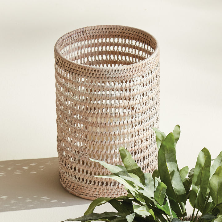 Large whitewash rattan hurricane on a pale console beside green branches, showing its open woven form in light, Transitional