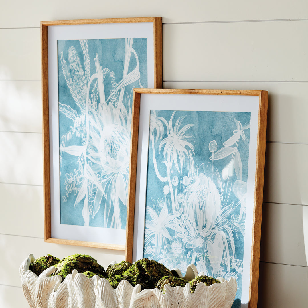 Two blue protea prints leaning against a wall above a textured planter, with white mats and wood frames visible. Coastal