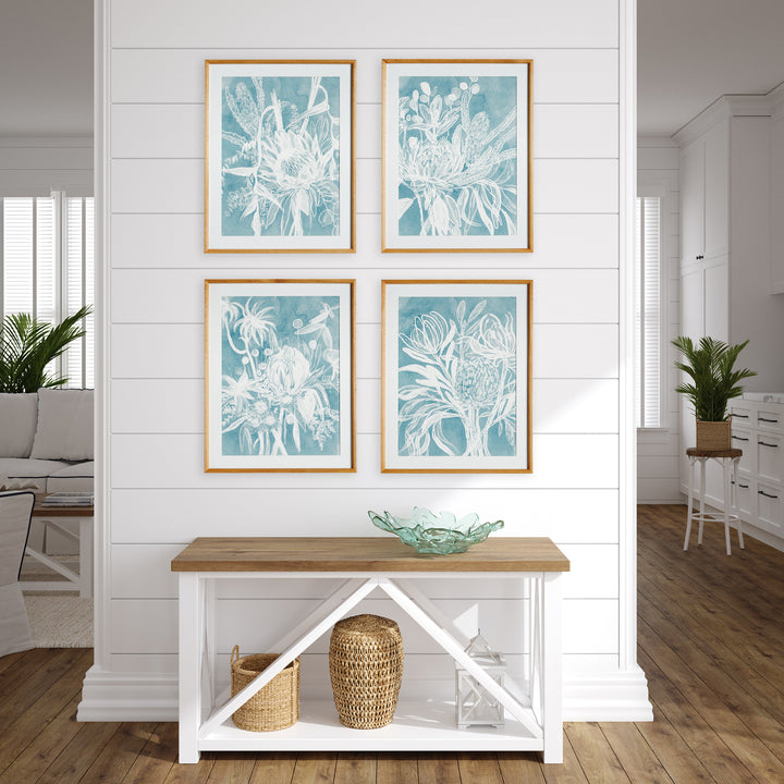 Four blue protea prints displayed in a wall grid above a console, with white mats and matching natural wood frames. Coastal