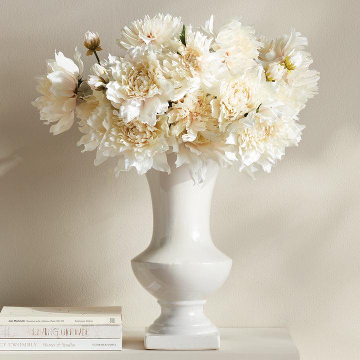 Large white ceramic trumpet vase holding pale flowers on a console beside a stack of books, arranged in Traditional style