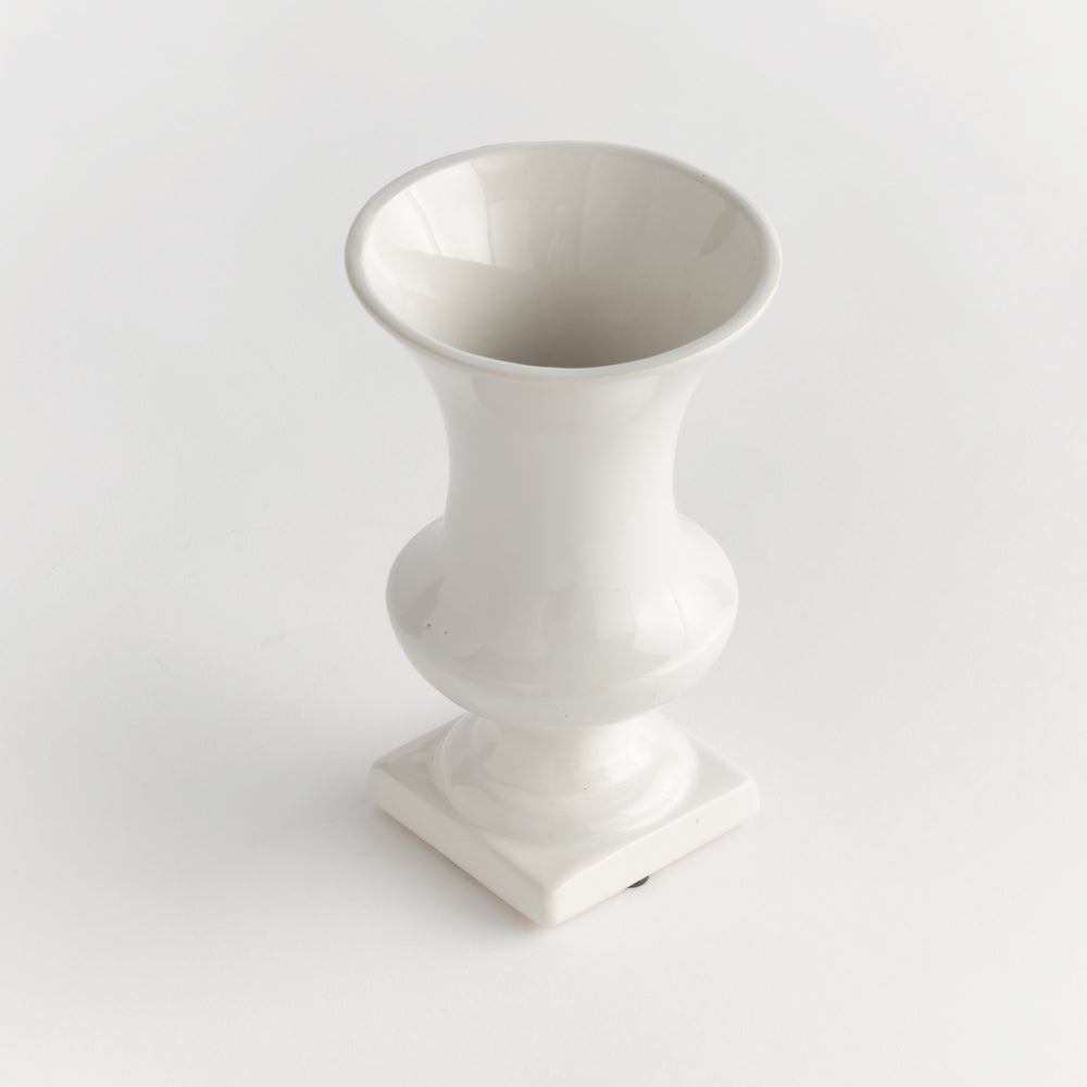 Angled view of a small white ceramic footed vase showing its open rim and shaped square base, presented in Traditional style
