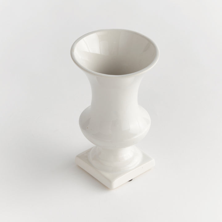Angled view of a small white ceramic footed vase showing its open rim and shaped square base, presented in Traditional style