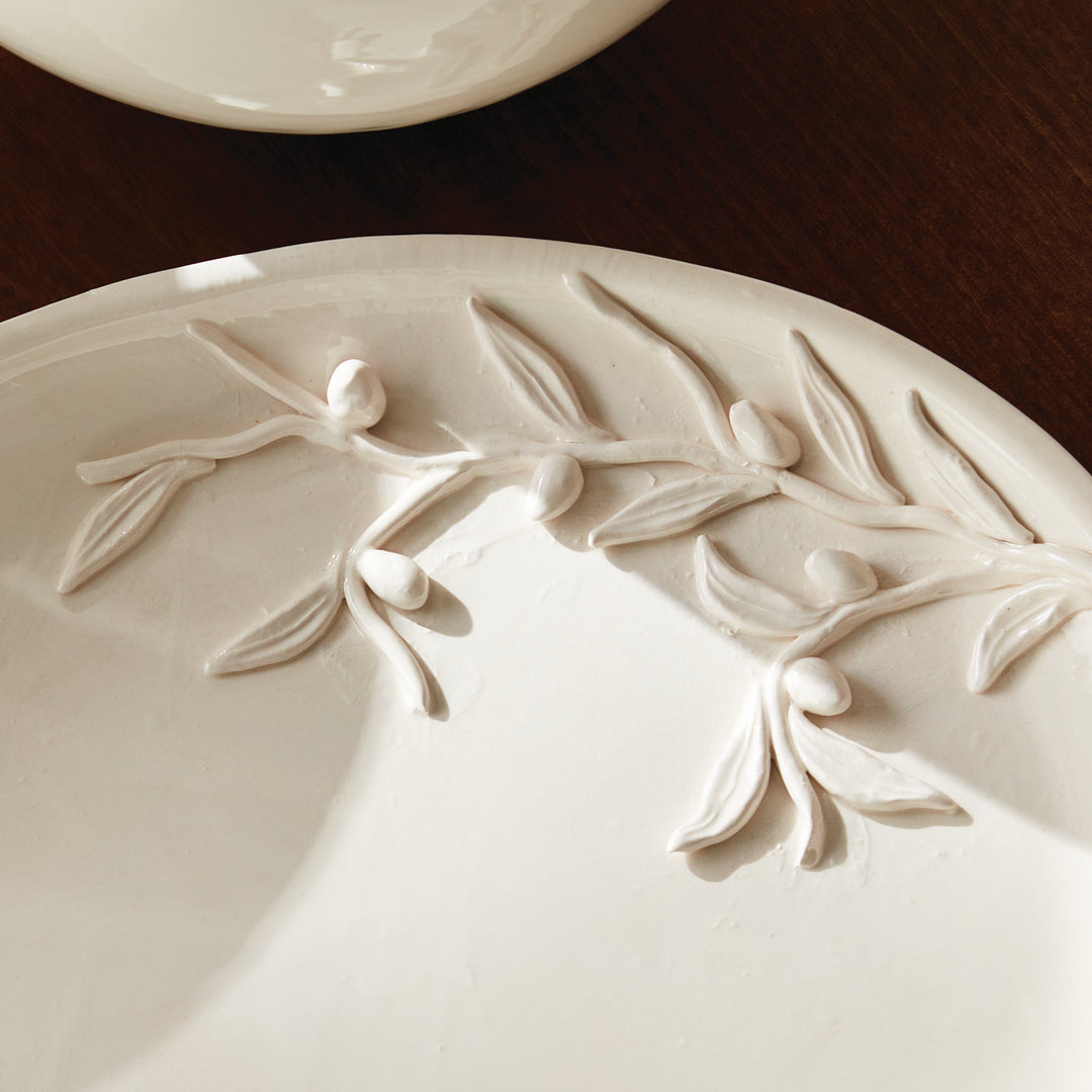 Close overhead view of olive leaves and fruit in ceramic relief across a softly glazed oval decorative tray. Traditional