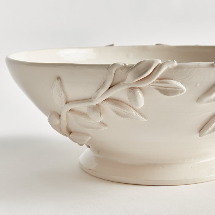 Close view of raised olive branches and leaves on a white glazed ceramic bowl, above its round pedestal base. Traditional