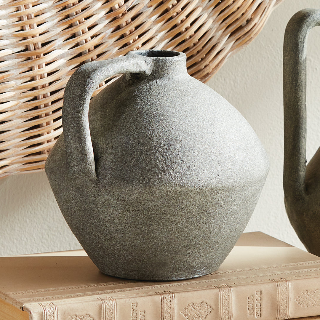 Gray terracotta rounded jug vase styled on books beside a taller jug, with woven wall decor in the background. Modern Organic