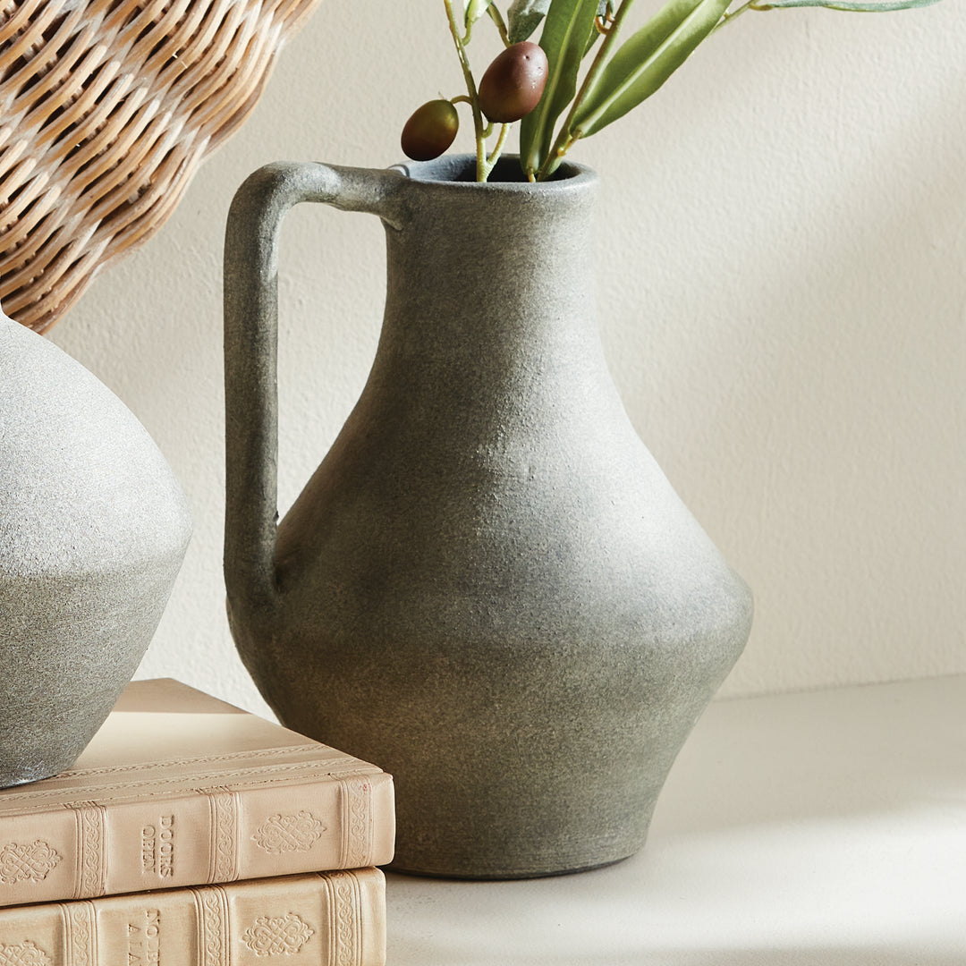 Gray terracotta long-neck jug vase styled with olive stems beside a rounded jug, books and woven wall decor. Modern Organic