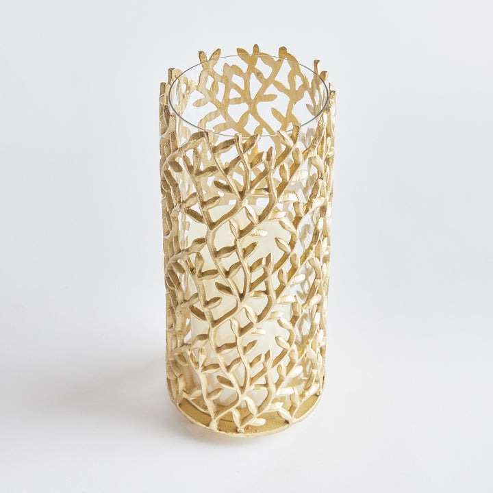 Cutout metal hurricane lantern in matte gold with leaf motif, perfect for modern botanical and nature-inspired decor themes.

