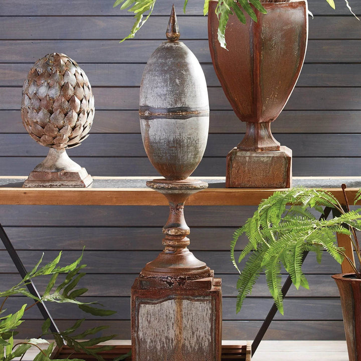 Oversized metal finial with rustic industrial finish styled on a wood shelf with antique accents and greenery.
