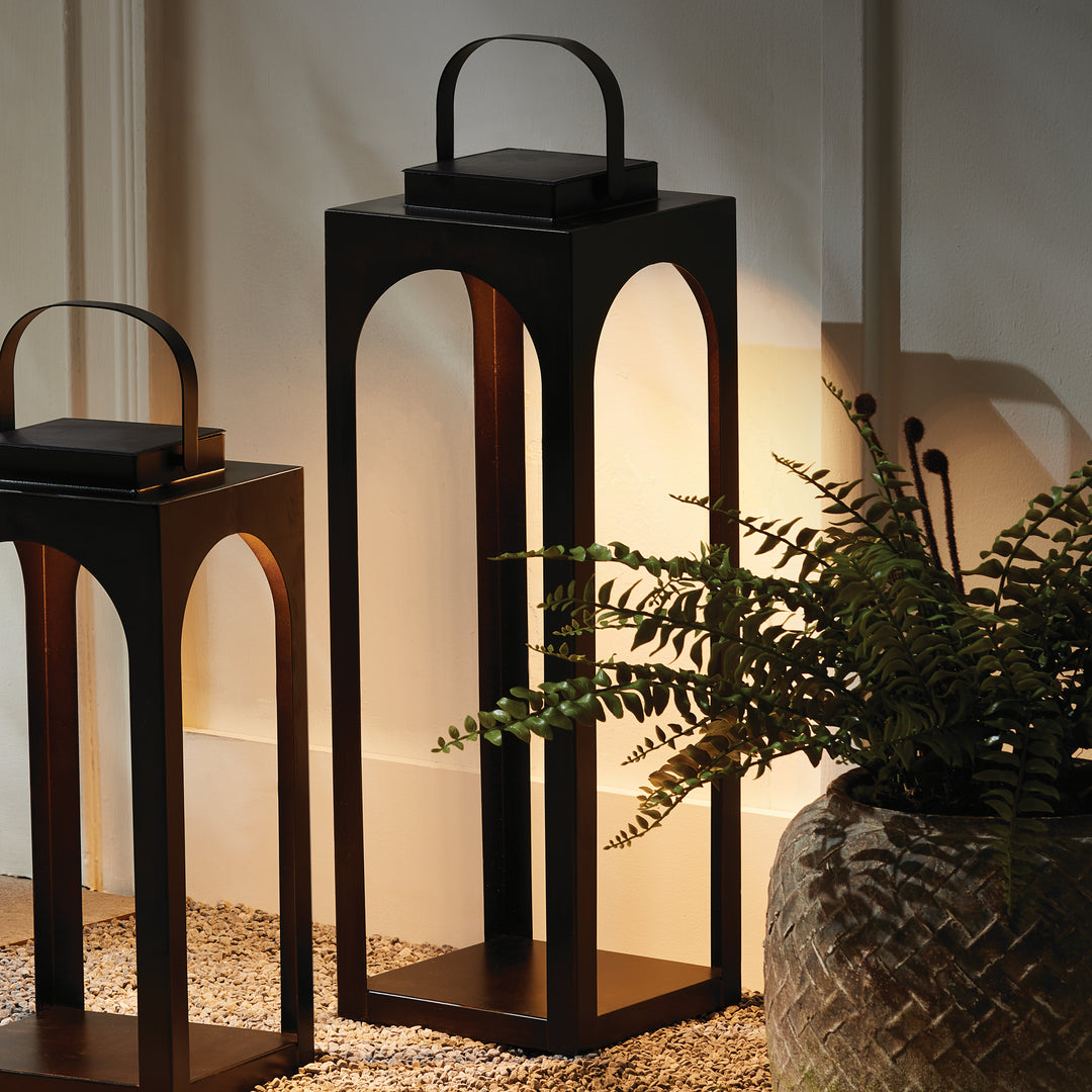 Pair of modern arched solar LED outdoor lanterns illuminating a covered patio with warm ambient light beside lush greenery – Transitional