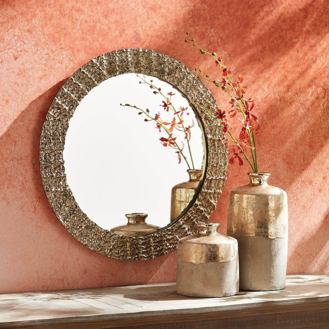 Modern round mirror styled on wood console with warm wall and metallic vases, showing gold textured frame