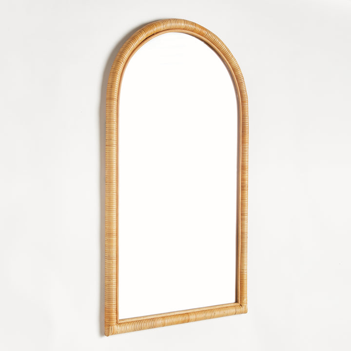 Natural rattan arched wall mirror with honey tan wrapped frame, shown at an angle against a simple white studio wall, Coastal