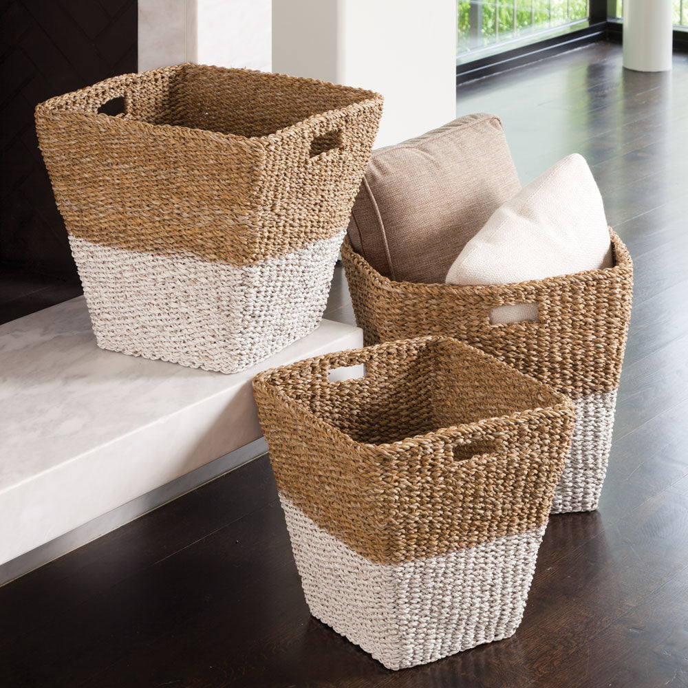 Set of three seagrass baskets styled with pillows near a marble fireplace in a modern neutral living space.