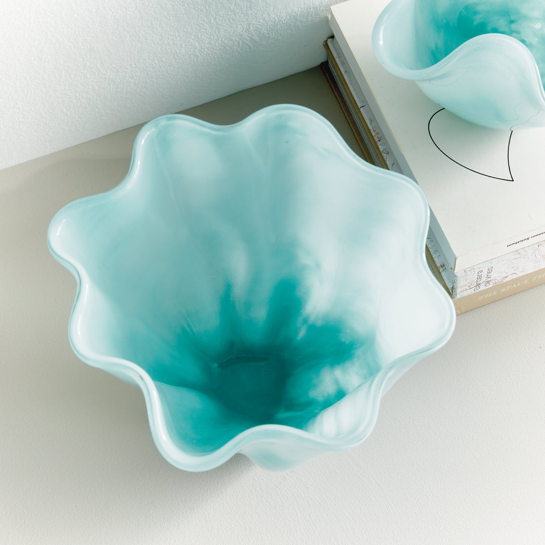 Overhead styled view into a light blue glass vase beside books, showing a waved rim and soft watercolor-like color. Coastal