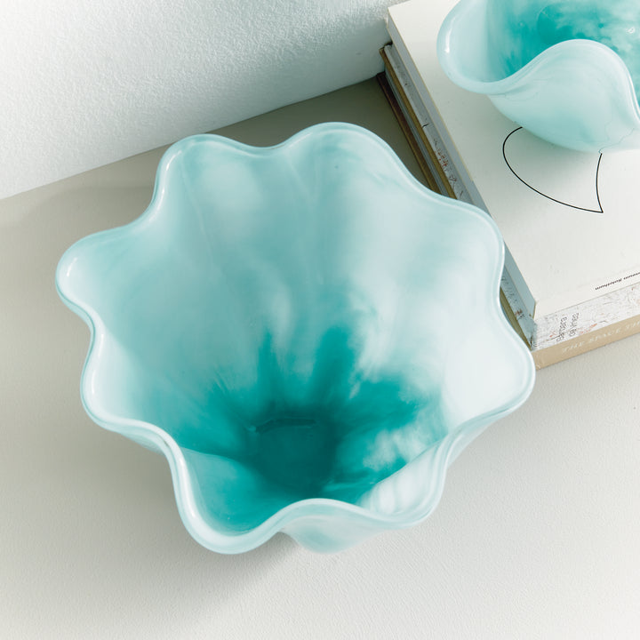 Overhead styled view into a light blue glass vase beside books, showing a waved rim and soft watercolor-like color. Coastal