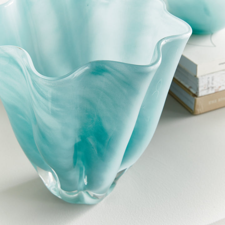 Close styled view of a light blue glass vase beside books, showing the wavy rim, flowing sides and translucent color. Coastal