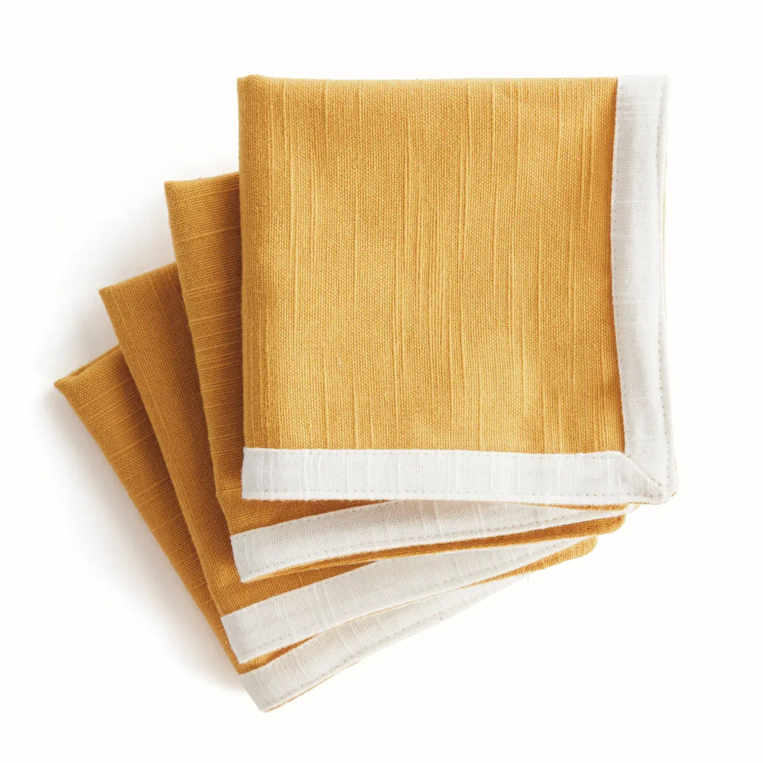 Ochre cotton cocktail napkins with white borders, folded in a stacked arrangement to show their woven texture, Transitional