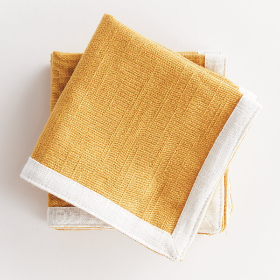 Folded ochre cotton cocktail napkins edged in white, layered at angles to show the square cloth and its borders, Transitional