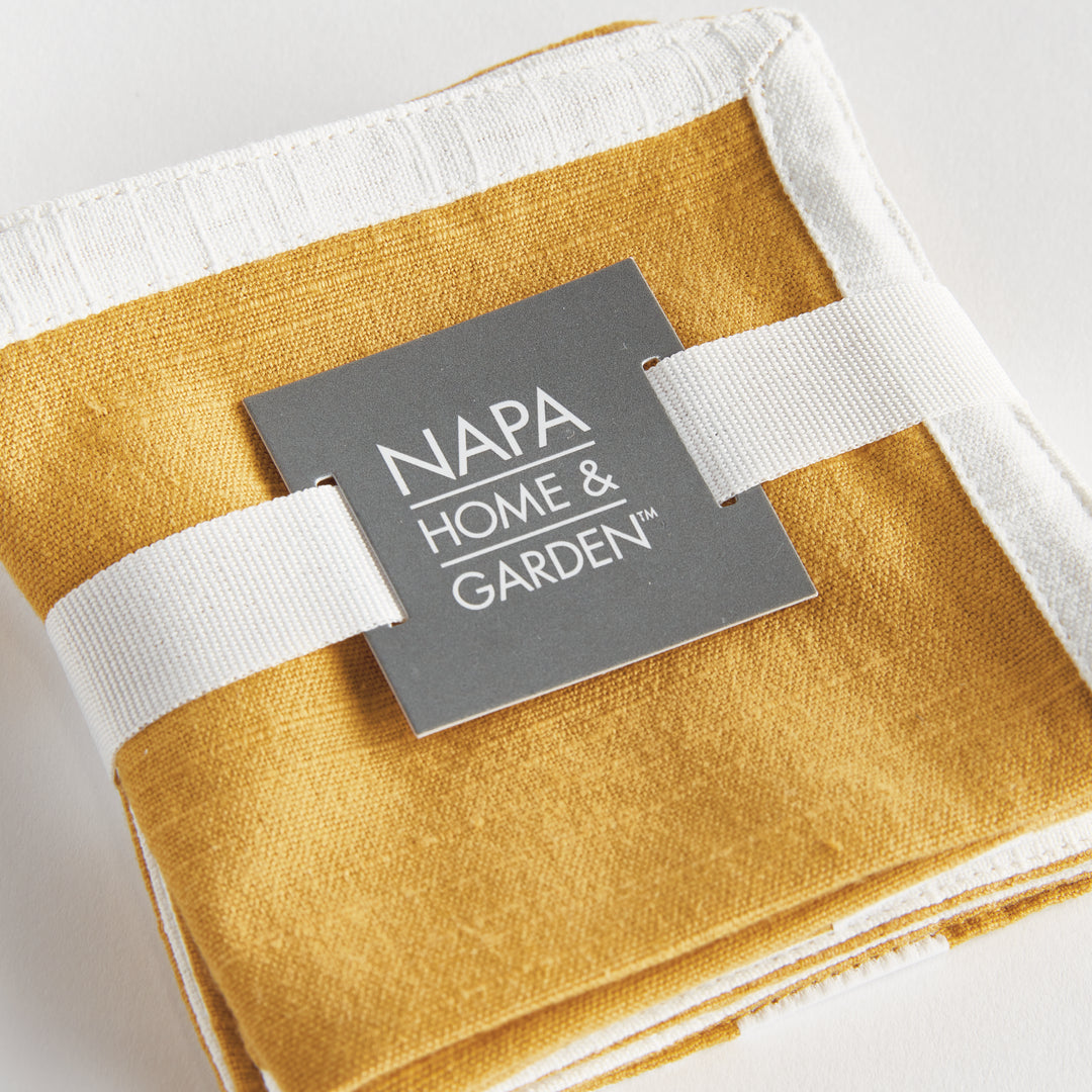 Close view of ribbon-tied ochre cotton cocktail napkins, showing white borders and the folded fabric on white, Transitional