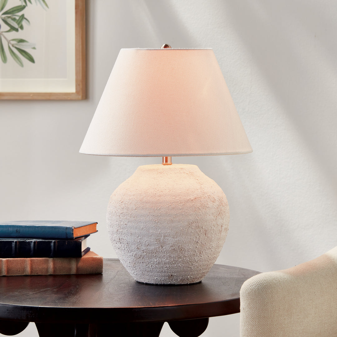 Orla ceramic table lamp glowing beside stacked books on a dark wood table with an upholstered chair nearby. Modern Organic
