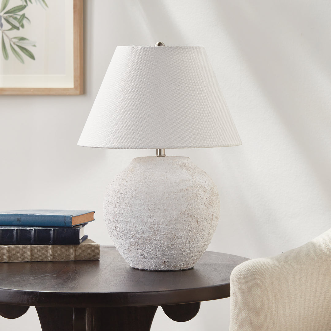 Orla white textured ceramic lamp with linen shade on a dark wood side table beside books and a fabric chair. Modern Organic
