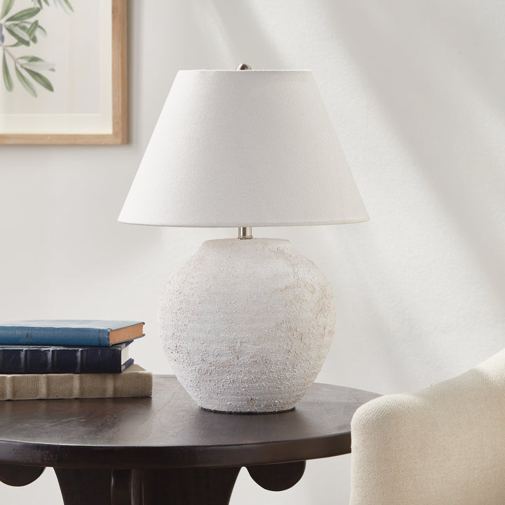 Orla white textured ceramic lamp with linen shade on a dark wood side table beside books and a fabric chair. Modern Organic