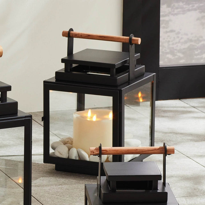 Medium black lantern styled outdoors with candle glowing on patio floor next to seating