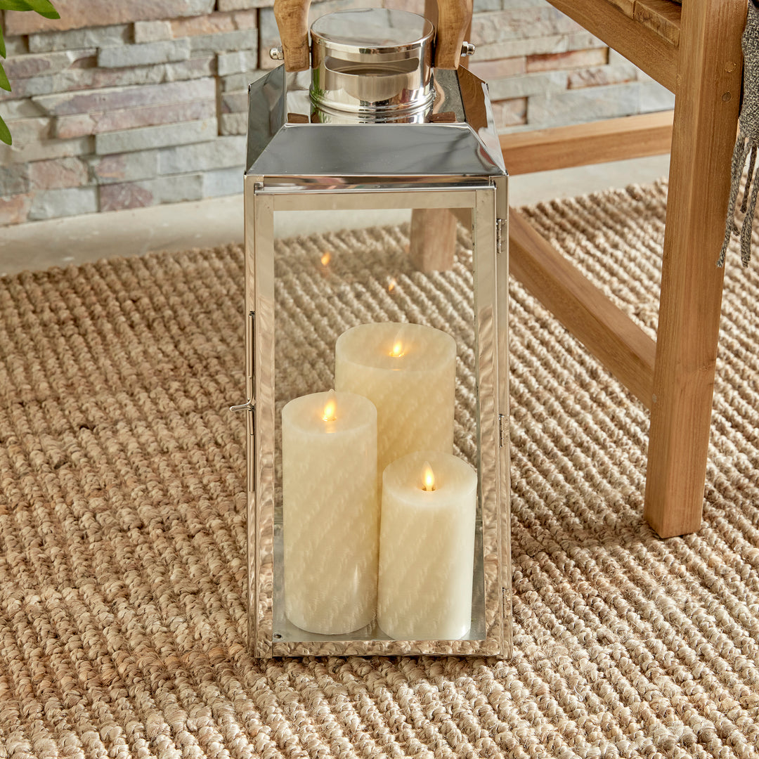 Small outdoor stainless steel lantern on woven rug near green plant with candles and wood chair coastal lantern