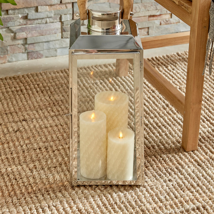 Small outdoor stainless steel lantern on woven rug near green plant with candles and wood chair coastal lantern