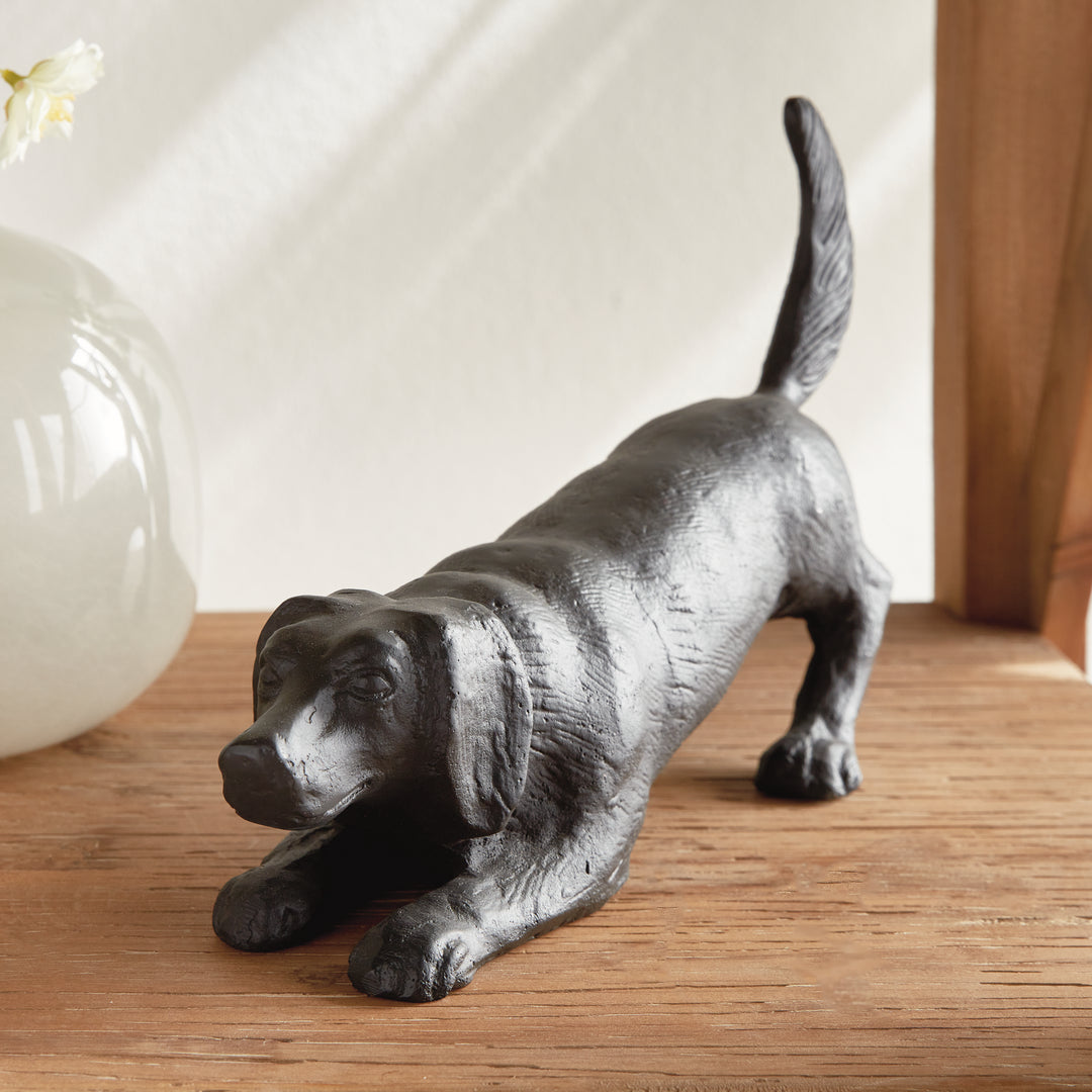 Black aluminum playful dog sculpture with bronze undertones photographed on a wooden tabletop in a styled interior setting.