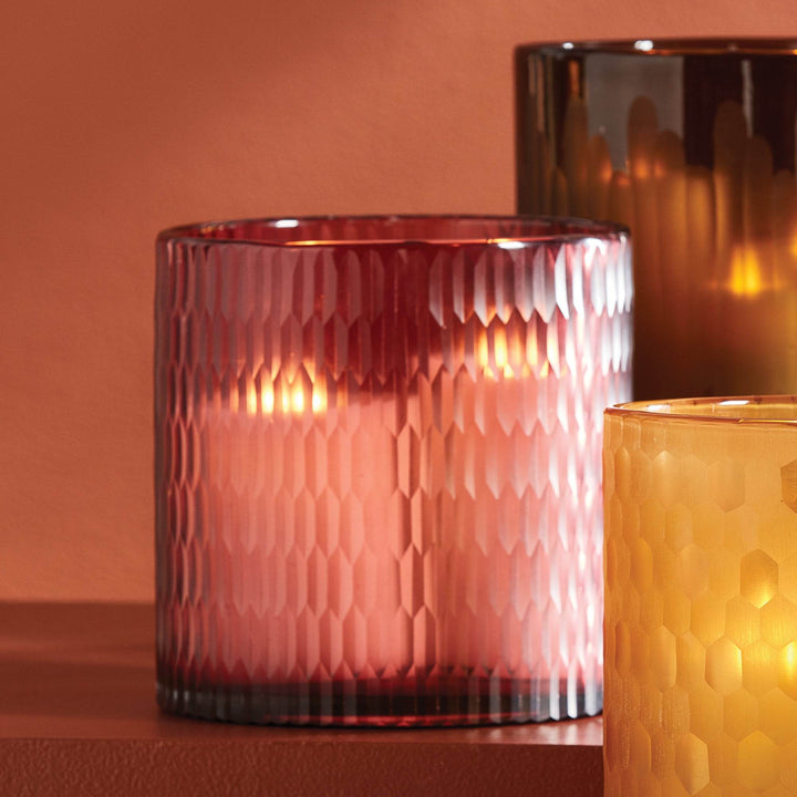 Plum glass hurricane styled with amber and brown glass vessels, showing warm candlelight and faceted surface texture – Modern Glam.

