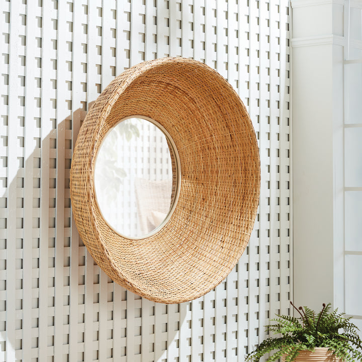 Rattan-framed wall mirror reflecting light on a sunlit white lattice wall with organic shadows

