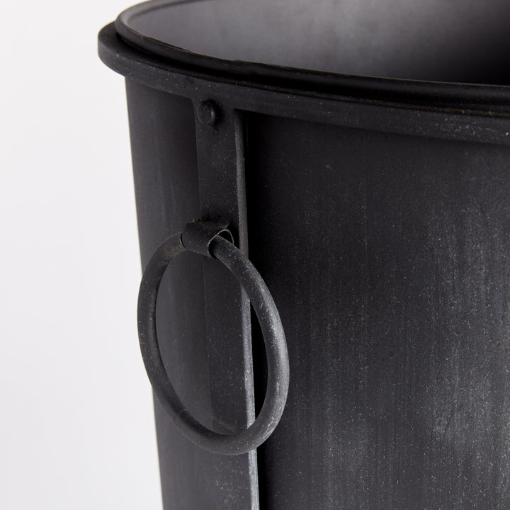 Large iron planter detail showing the ring handle, rolled rim and aged zinc surface beside a frame support. Transitional