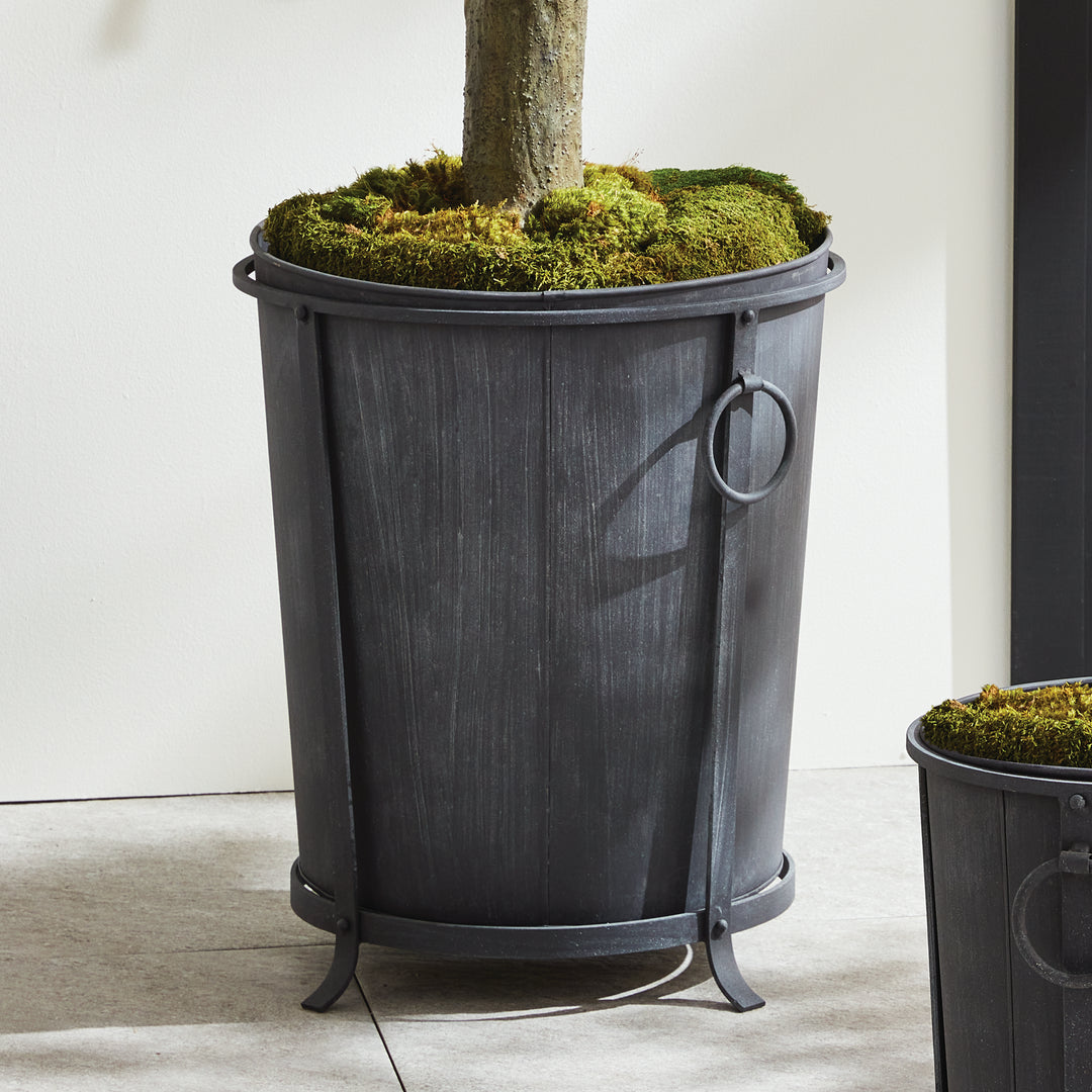 Large iron planter styled with a tree and moss at the rim, showing its ring handles and raised round frame. Transitional