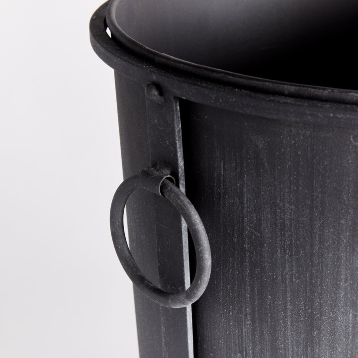 Medium iron planter detail showing the ring handle, rolled rim and aged zinc surface beside a frame support. Transitional
