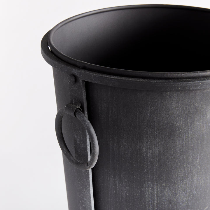 Small iron planter detail showing the ring handle, rolled rim and aged zinc surface beside a frame support. Transitional