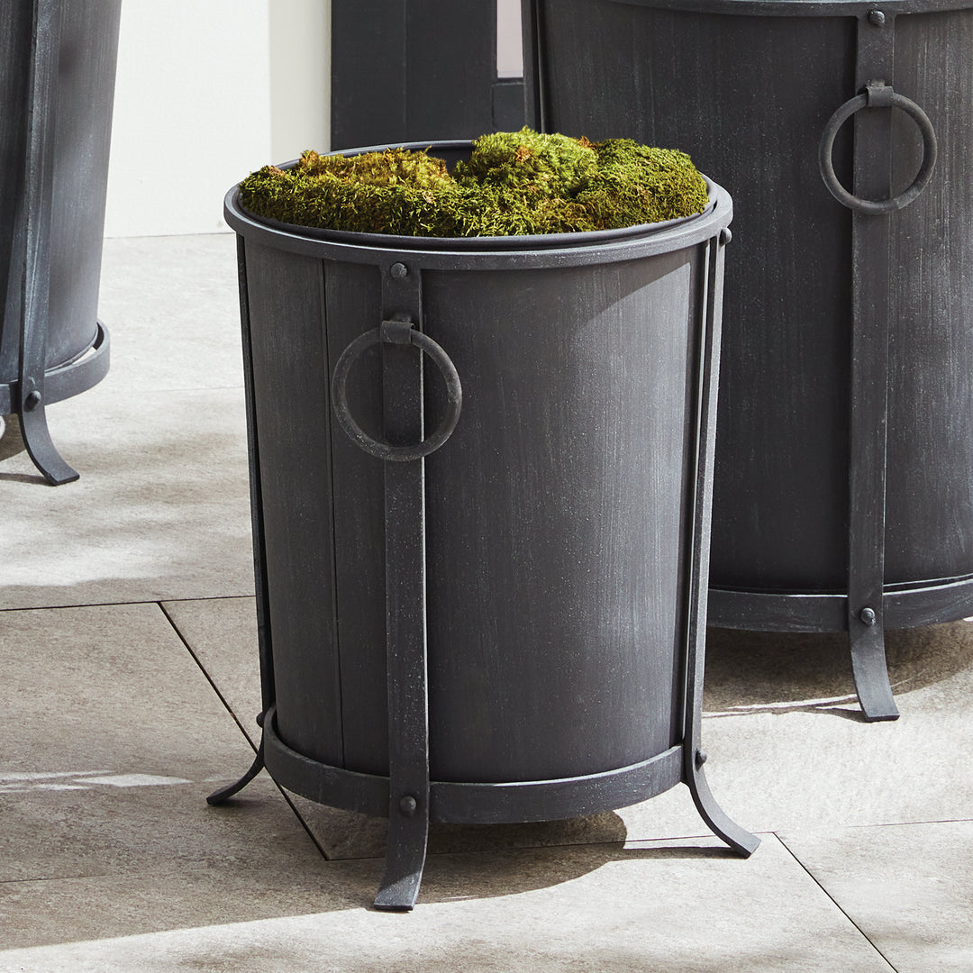 Small iron planter styled with a tree and moss at the rim, showing its ring handles and raised round frame. Transitional