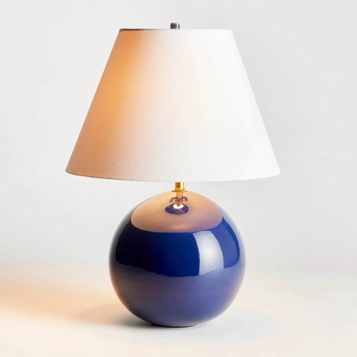 Illuminated blue glazed ceramic table lamp with a glowing linen shade and rounded glossy base on a pale surface. Transitional