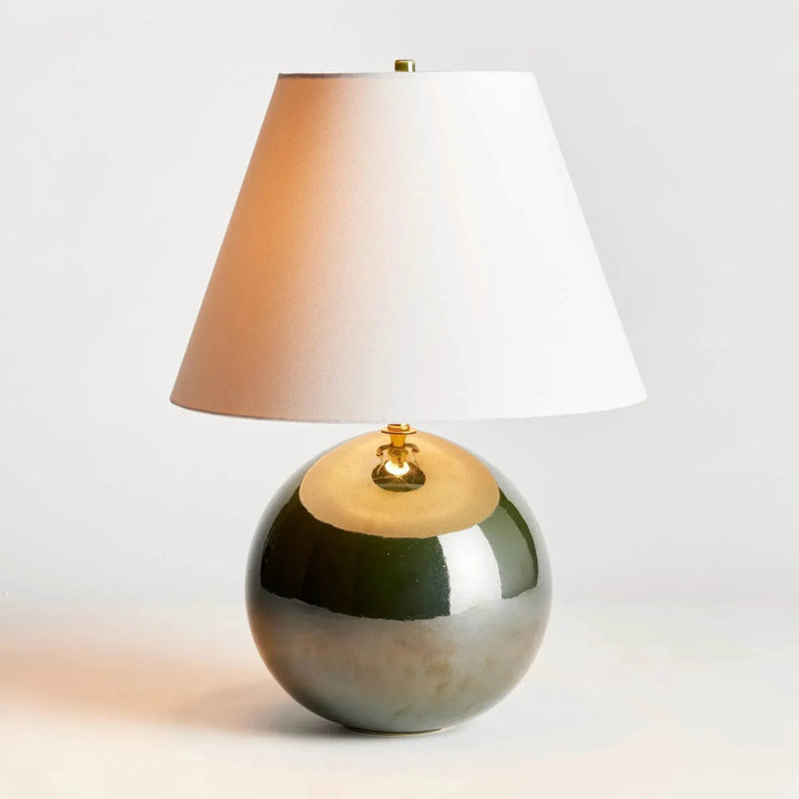 Illuminated green ceramic table lamp with a warm glowing linen shade and rounded glossy base on a pale surface. Transitional
