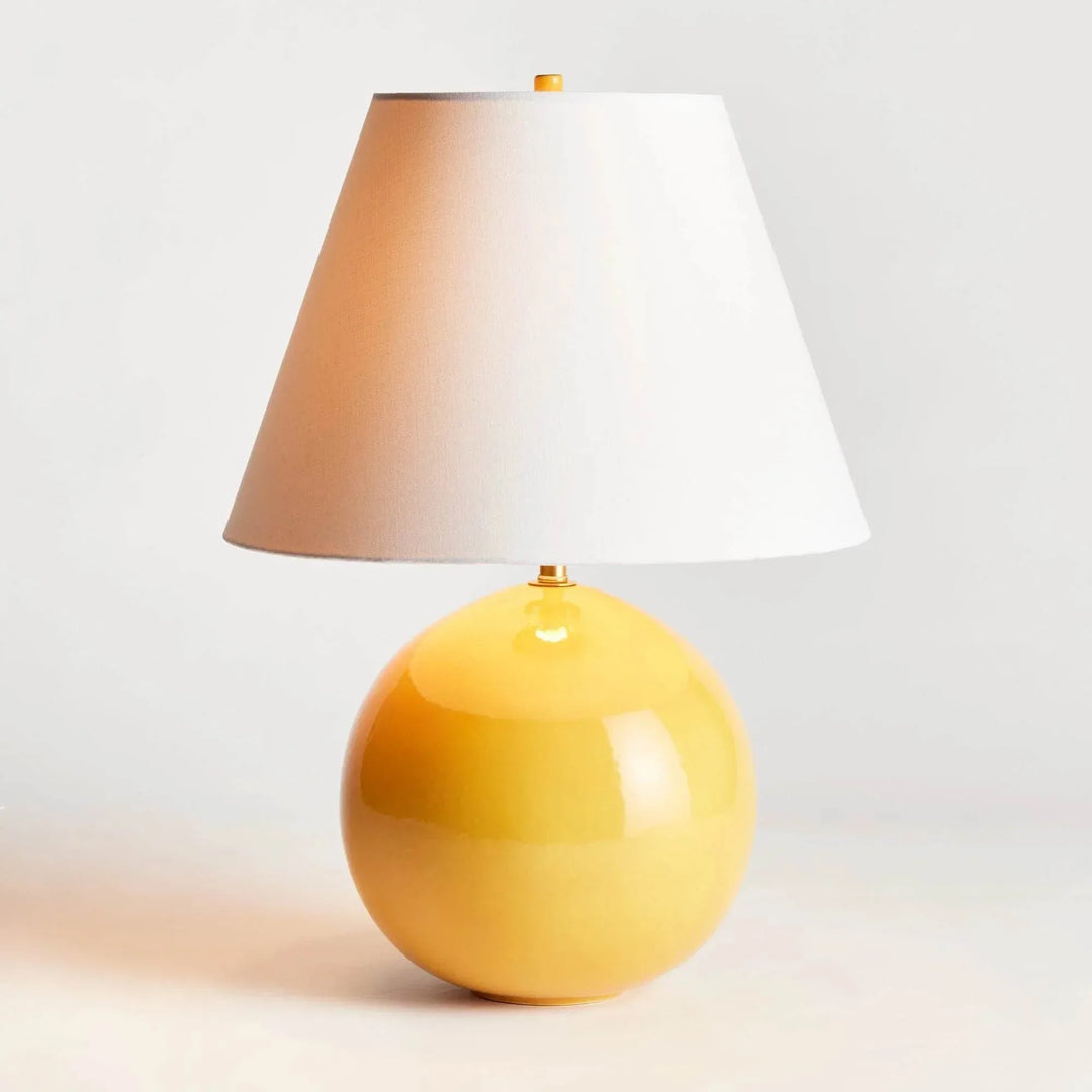 Illuminated marigold ceramic table lamp with a glowing linen shade and rounded glossy base on a pale surface. Transitional