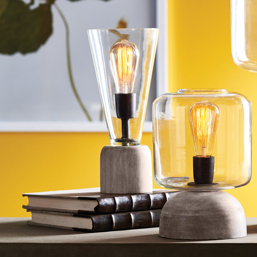 Tapered glass accent lamp on dark stacked books beside a second rounded glass lamp, with both bulbs glowing - Transitional