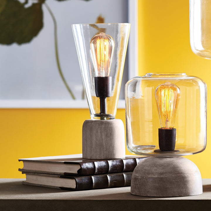 Tapered glass accent lamp on dark stacked books beside a second rounded glass lamp, with both bulbs glowing - Transitional