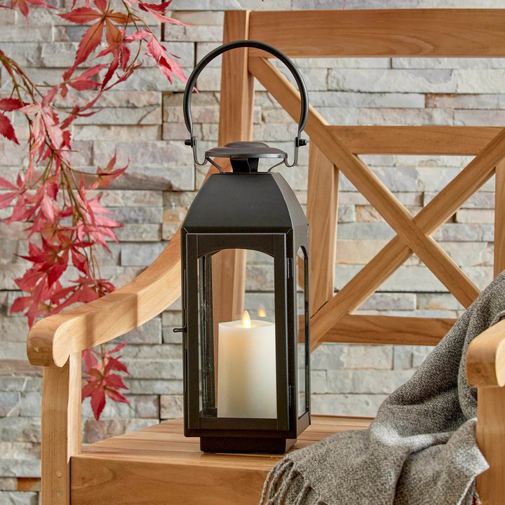 Small black outdoor lantern with pillar candle on wooden bench against stone wall; warm, welcoming outdoor vignette wide view – Transitional Patio Decor
