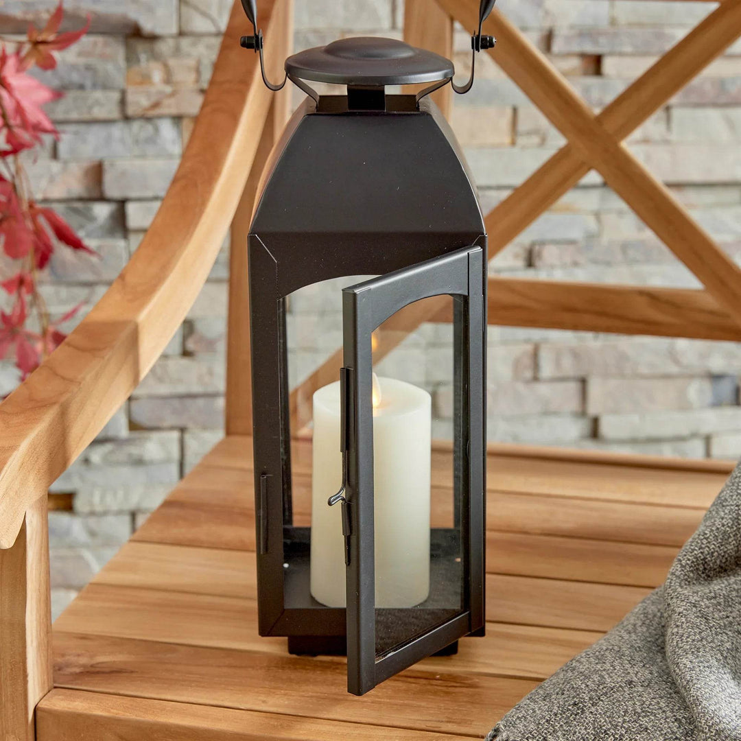 Small black outdoor lantern on bench with front door open and pillar candle visible; latch detail and glass panels shown – Transitional Lighting