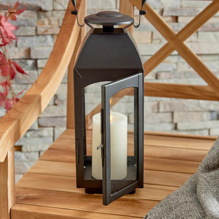 Small black outdoor lantern on bench with front door open and pillar candle visible; latch detail and glass panels shown – Transitional Lighting