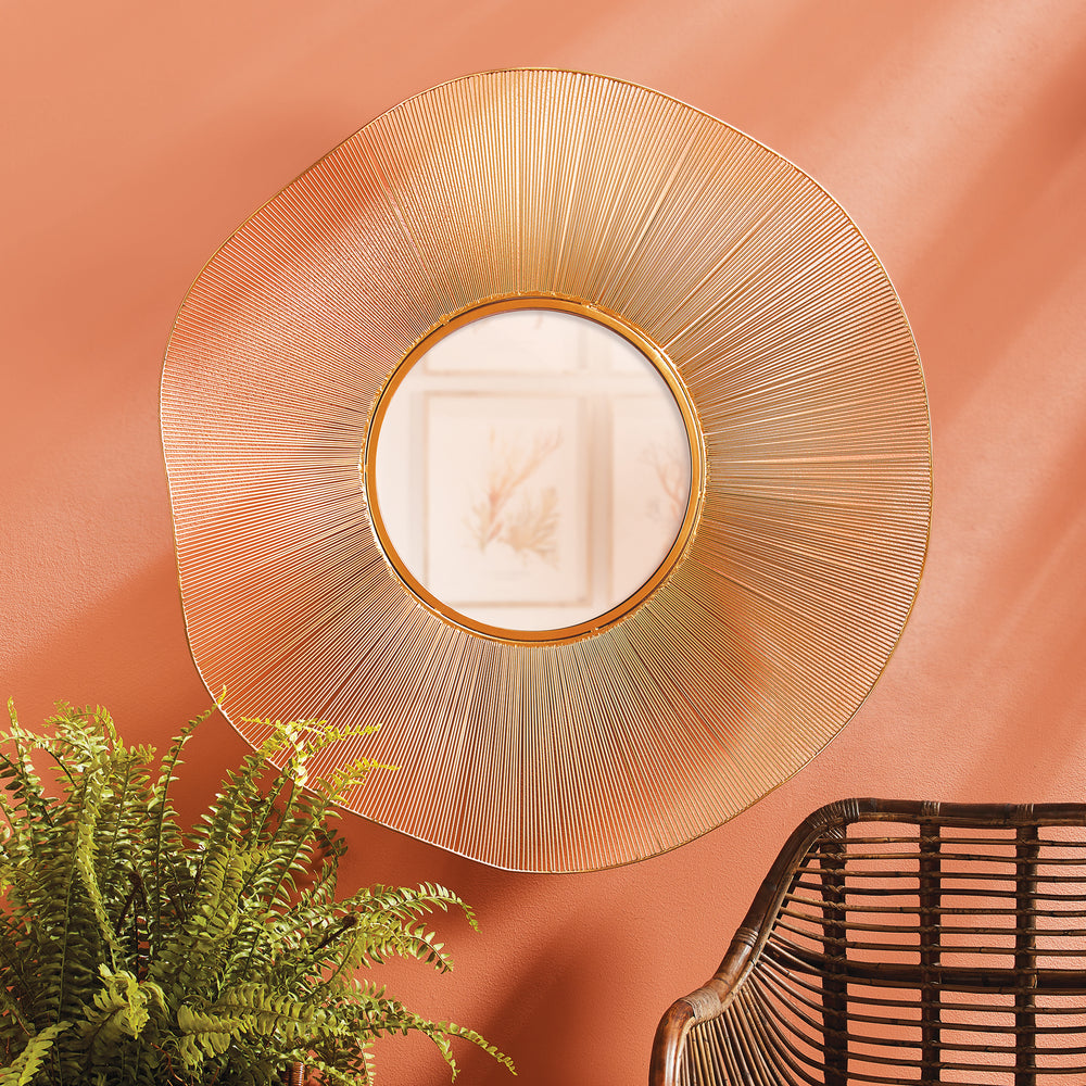 Gold openwork round wall mirror on a warm coral wall, styled beside leafy green foliage and dark woven seating - Organic Glam