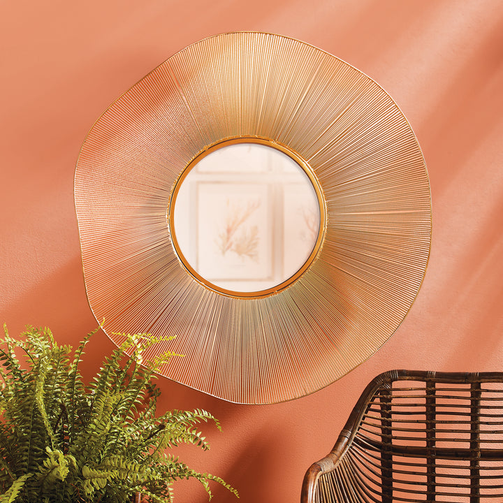 Gold openwork round wall mirror on a warm coral wall, styled beside leafy green foliage and dark woven seating - Organic Glam